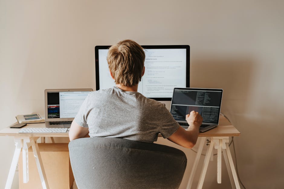 A person with short, light brown hair is seated at a light wooden desk, working on a laptop with a black keyboard and screen displaying coding or text documents. In front of them is a larger desktop m