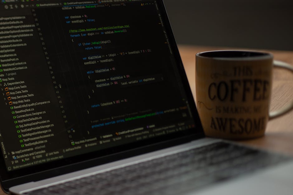 A person with short, light brown hair is seated at a light wooden desk, working on a laptop with a black keyboard and screen displaying coding or text documents. In front of them is a larger desktop m